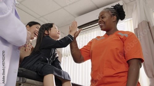 Happy Asian girl giving a high five to African female nurse after treatment in hospital, with her mother and doctor smiling nearby.