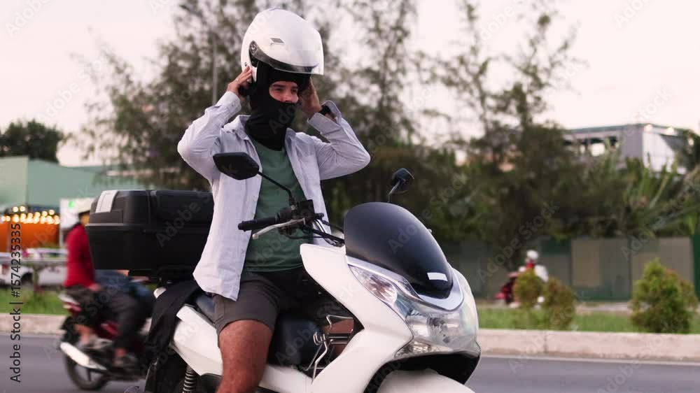 The man puts on his helmet while sitting on the moped. The man puts on his helmet while sitting on the moped.