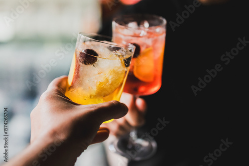 Two hands clinking cocktail glasses garnished with fruit and ice, celebrating with colorful drinks in focus and a blurred festive background.