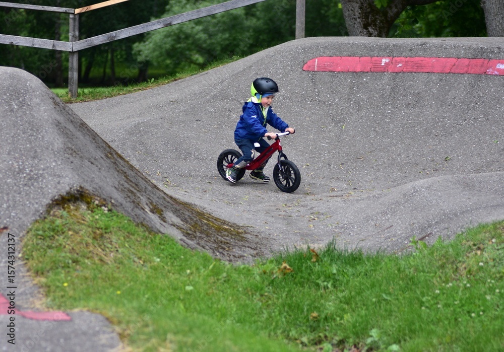 Obraz premium Active young boy in blue jacket and helmet riding a red balance bike on an outdoor pump track. Child practicing cycling skills in green park on summer day. Concept of active childhood and sport.
