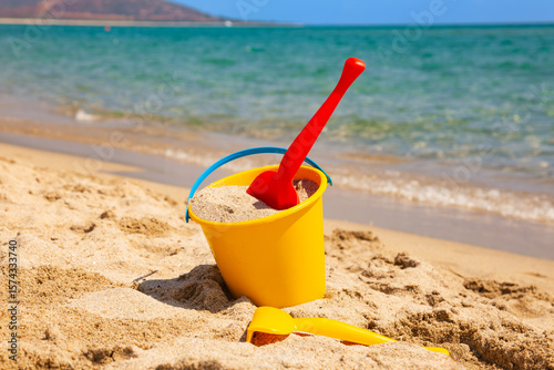 Yellow bucket with red plastic shovel filled with sand on sunny beach