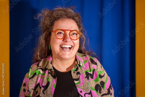Cheerful female designer with curly hair, orange glasses, laughing while glancing sideways against vibrant blue and yellow backdrop