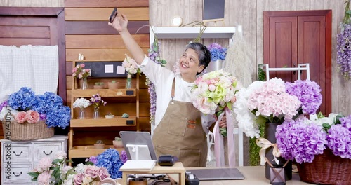 Asian elderly female florist smiling while holding bouquet and taking selfie in flower shop, capturing joyful moment as small business owner celebrating floral creation after retirement