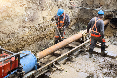 Three utility workers installing a pre-insulated steel pipe at a district heating construction site. Precise alignment and teamwork during underground pipe assembly for thermal energy distribution.
