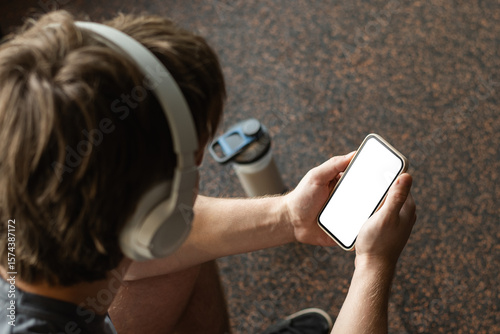 Young sporty man and bodybuilder holding smartphone with empty white screen, while taking break between exercises at gym. Mock up. Close up.