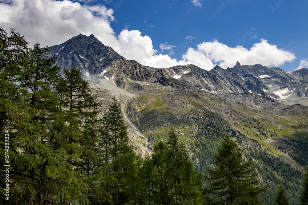 Obraz premium Scenic view of rugged alpine mountains in Switzerland with rocky peaks, green slopes, and scattered trees under a blue sky with white clouds.