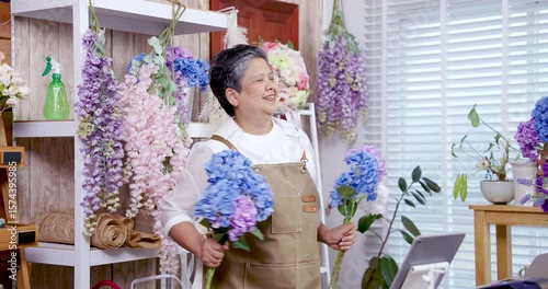 Asian senior woman florist dancing while hugging bouquet of blue flowers smiling eyes closed slowmotion capturing emotion joy of independent creative business in cozy floral workspace