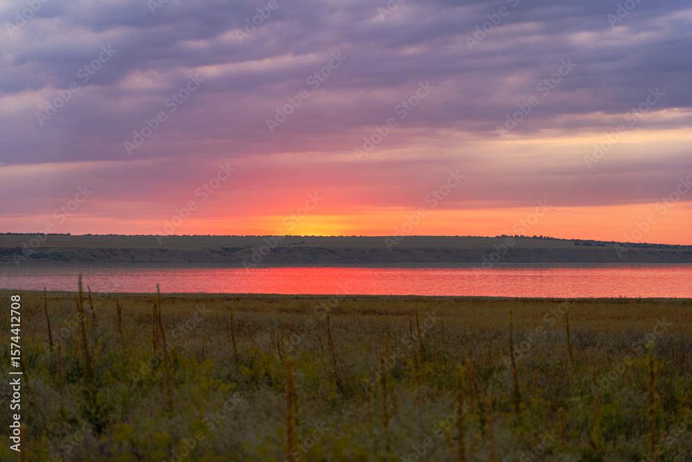 Obraz premium Colorful summer sunset over river with cloudy sky and meadow in foreground, dramatic warm light reflecting in the water near coastal hills