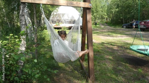 Girl relaxing in hammock. View of cute teenage girl in white t-shirt swinging in hammock at forest camping. Teenager using smartphone.