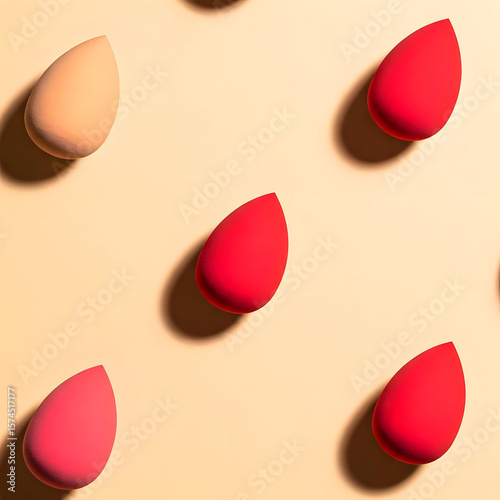 A top-view fashion flat lay featuring teardrop-shaped beauty blenders in red and nude tones, forming a repeating triangle pattern on a soft beige surface