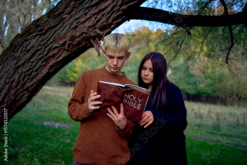 a 15-year-old teenage couple in love reading the holy bible together in the park on a cold autumn evening