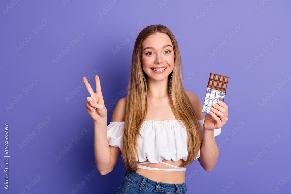 Obraz premium Charming young woman smiling and posing with a chocolate bar on a vibrant purple background in a summer outfit