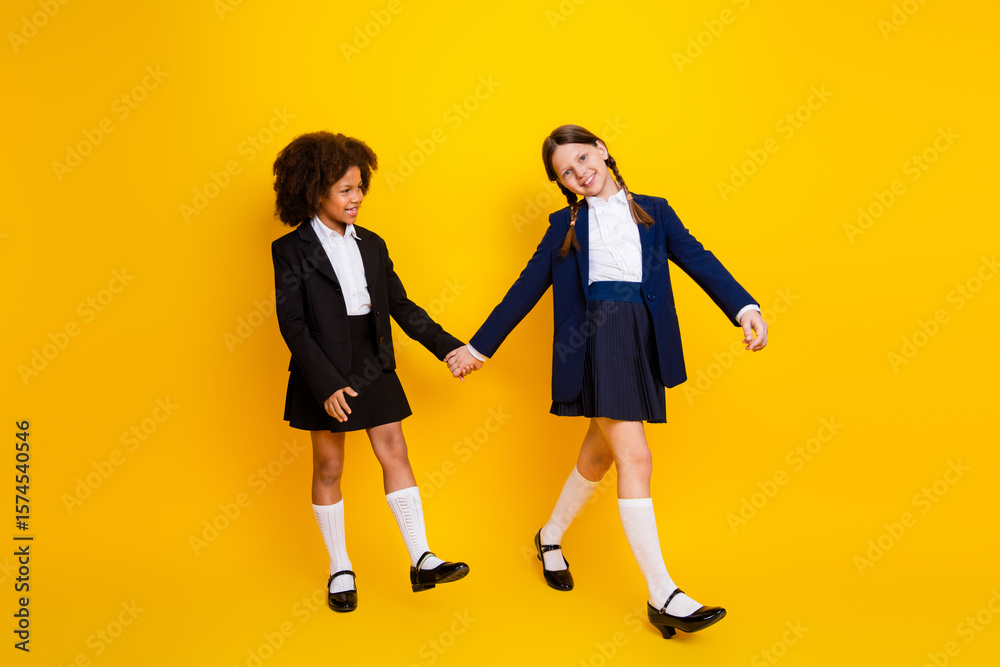 Fototapeta premium Happy schoolgirls holding hands on vibrant yellow background showcasing friendship and diversity in stylish school uniforms