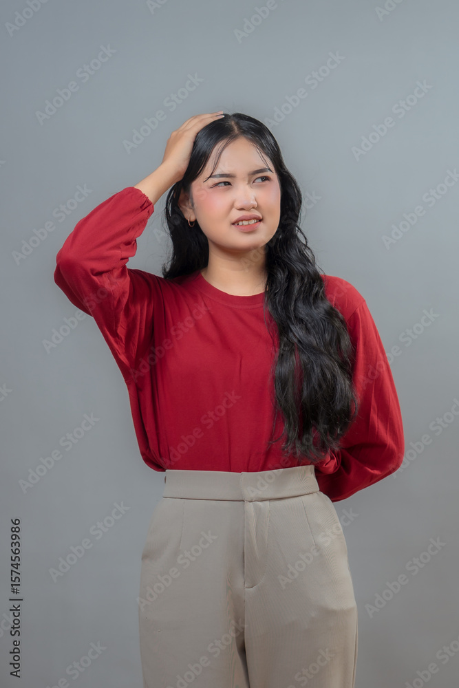 A thoughtful Indonesian woman in a red top and beige pants scratching her head with a confused expression. The photo captures a moment of uncertainty or contemplation. A thoughtful Indonesian woman in a red top and beige pants scratching her head with a confused expression. The photo captures a moment of uncertainty or contemplation.