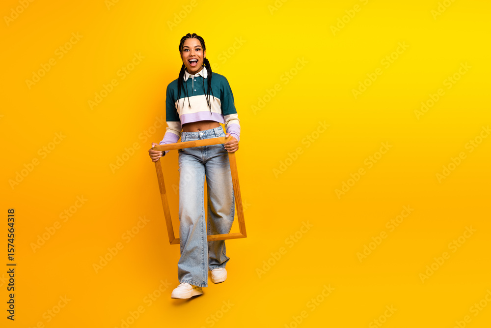 Fototapeta premium Smiling young woman in casual outfit holding wooden frame on vibrant yellow background, showcasing stylish and trendy look