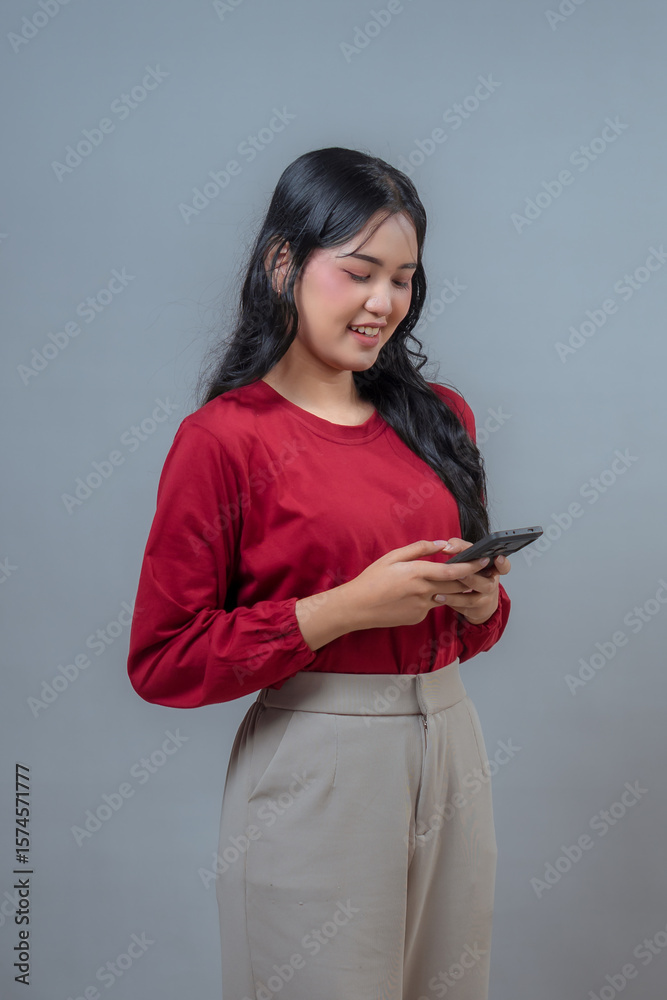 Smiling Indonesian woman in red blouse and beige trousers uses smartphone, standing sideways on gray backdrop. Relaxed posture and delighted look capture a modern digital lifestyle. Smiling Indonesian woman in red blouse and beige trousers uses smartphone, standing sideways on gray backdrop. Relaxed posture and delighted look capture a modern digital lifestyle.