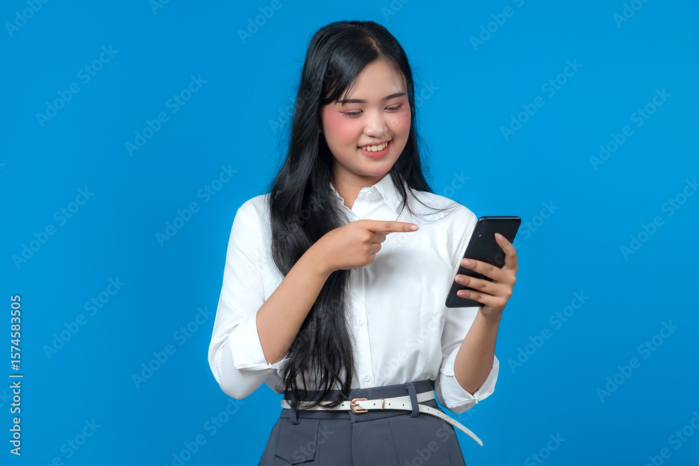 Smiling young Asian woman in a white shirt pointing at her smartphone screen while looking at it with interest, standing against a blue background, promoting or showing information. Smiling young Asian woman in a white shirt pointing at her smartphone screen while looking at it with interest, standing against a blue background, promoting or showing information.