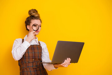 Young woman in stylish attire inspecting laptop closely with magnifying glass against bright yellow background