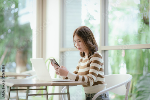 Fotografía A young woman sits in a bright cafe, focused on her smartphone, with a laptop open in front of her, enjoying a peaceful, productive moment