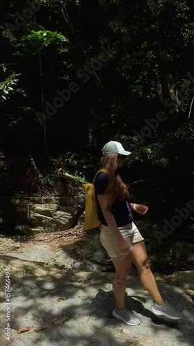 Vertical video. Woman with Yellow Backpack Walking on Sunlit Rocks in Jungle Clearing, Side View, Nature Travel Concept