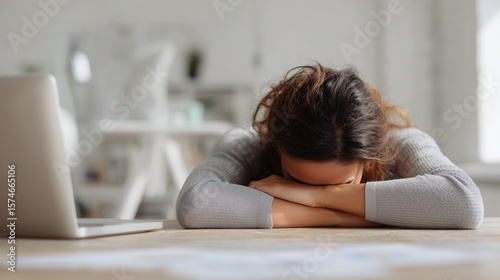 Woman resting head on folded arms at desk with laptop, showing tiredness and exhaustion in bright office environment