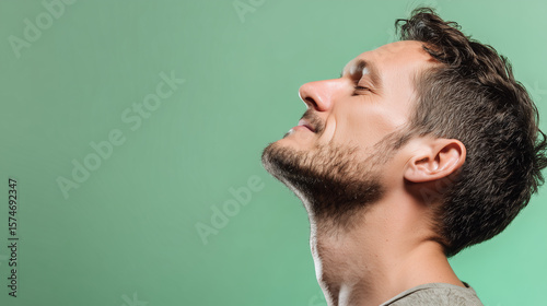 Man with beard closing eyes and exhaling deeply with relaxed expression against green background, showing calmness and stress relief in close up side profile portrait