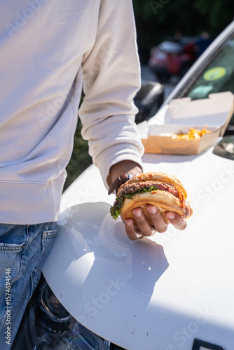 Foto Homme acoudé à une voiture tenant un burger dans sa main, capturé en extérieur dans une ambiance décontractée