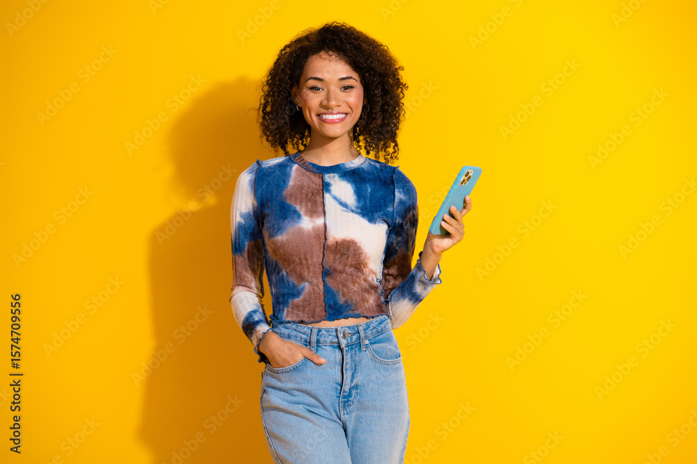 Fototapeta premium Smiling young woman with smartphone against vibrant yellow background wearing trendy casual outfit with curly hair