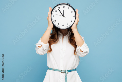 Fotografie Young successful employee business woman corporate lawyer wear shirt classic formal clothes work in office hold in hand cover eye with clock isolated on pastel light blue background studio portrait
