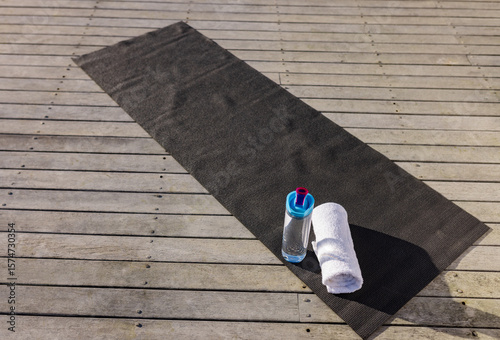 Black mat lying diagonally on wooden deck with water bottle, blue cap, pink spout, white towel