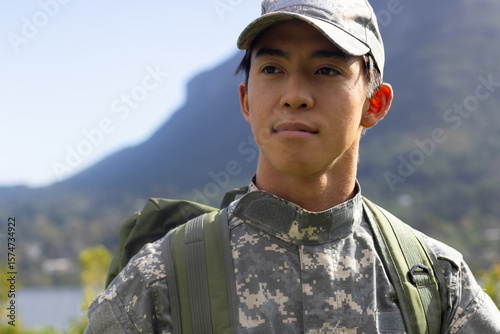 Asian male soldier in camo cap standing by lake near mountains carrying green backpack, copy space