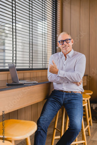 Successful senior manager showing thumbs up gesture while sitting on a stool in a modern office with laptop on counter, expressing satisfaction and approval