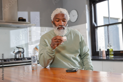 Senior African American man examining pill pack at kitchen island, glancing at smartphone and water
