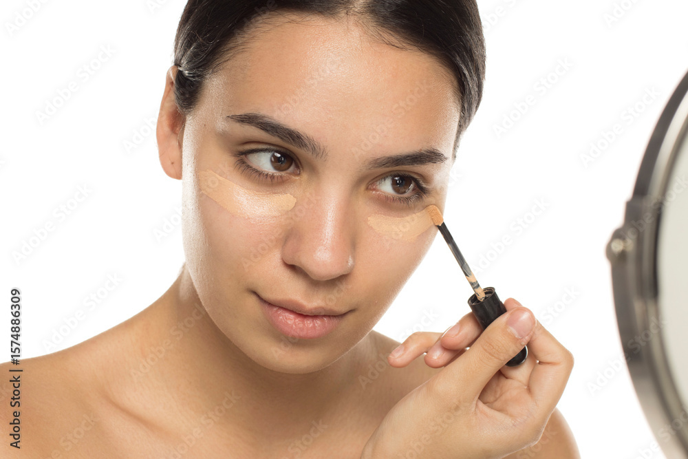 Young woman applying concealer under both eyes with applicator, looking in mirror, studio shot on white background. Young woman applying concealer under both eyes with applicator, looking in mirror, studio shot on white background.
