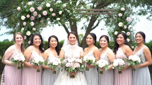 Bride and bridesmaids in dresses holding bouquets posing under floral archway smiling outdoors