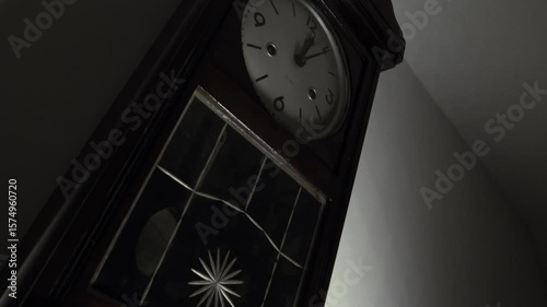 Dramatic low-angle, Dutch tilt zoom-in on a vintage pendulum wall clock. The quick movement of the hands suggests a time-lapse, symbolizing the passage of time. The moody, low-key lighting