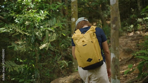 Woman in Cap and Yellow Backpack Hiking Up Rocky Forest Trail on Samui Island Surrounded by Dense Greenery, Rear Angle, Concept of Determination and Nature Exploration