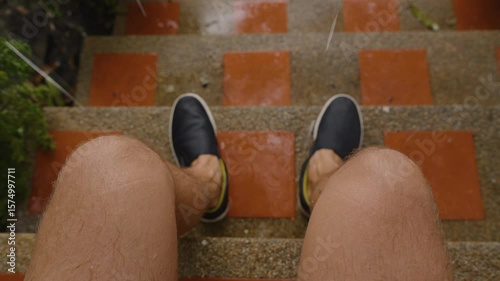 Wet Man Sitting on Brick Steps During Tropical Rain with Legs Visible from First Person Angle, Raindrops Falling on Red Tiles, First-Person View, Concept of Rain and Waiting