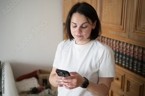 Surprised young woman in a room, looking at her phone opposite a bookshelf. Concept: unexpected news, digital connection, curiosity, modern life moments.