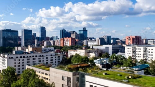 An aerial view of a city under a blue, cloudy sky. Modern and older buildings intermingle amidst green trees. The architecture varies