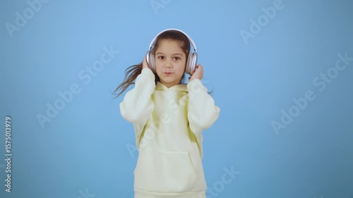 Child enjoys music while wearing headphones against a light blue background, smiling and expressing happiness with relaxed body language