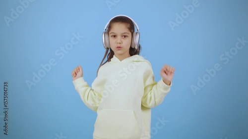 Child in light yellow hoodie dances joyfully with headphones against a blue background, showcasing a lively moment of music enjoyment and playful energy