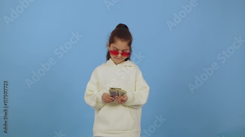 Child joyfully tossing money while wearing red sunglasses against a bright blue background, capturing a playful moment of fun and excitement