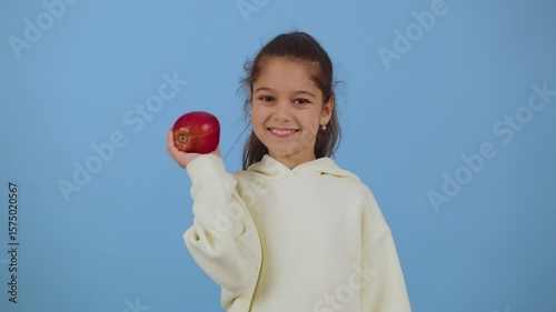 Young girl smiling while holding a red apple against a blue background during a playful indoor moment