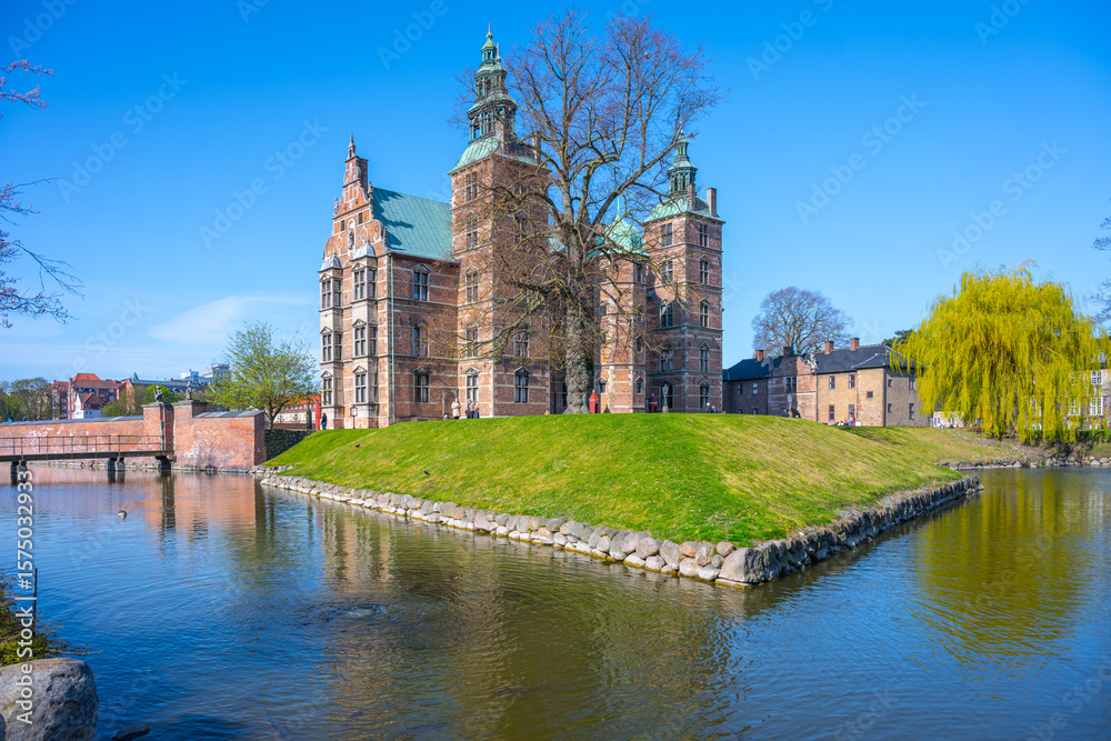 Obraz premium Rosenborg Castle showcases its stunning renaissance architecture in Copenhagen, surrounded by tranquil waters and lush greenery on a bright, clear day with blue skies.