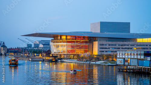 Copenhagen Opera House illuminates the evening sky with vibrant colors, reflecting beautifully on the water. Boats glide by as the cityscape provides a stunning backdrop in Denmark.