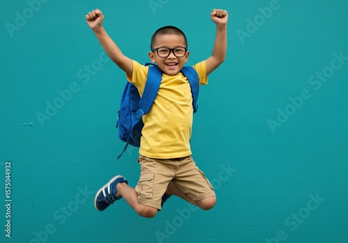 A young Asian boy celebrates with a joyful jump, wearing glasses and a backpack.