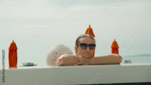 Woman with sunglasses rests arms on edge of pool facing camera with umbrellas and ocean behind, wide angle, Beachfront Resort Mood Concept