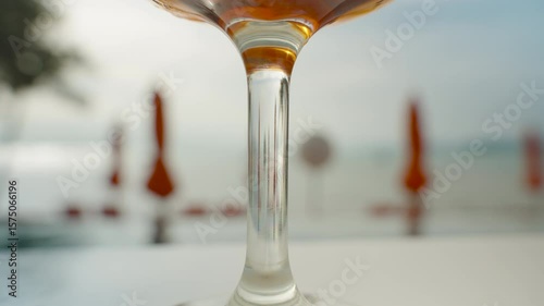 Close up of cocktail glass stem with blurred beach umbrellas and sea in background, shallow depth of field, Vacation Vibes Concept
