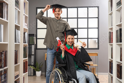 Female graduate in wheelchair with diploma and her classmate at library